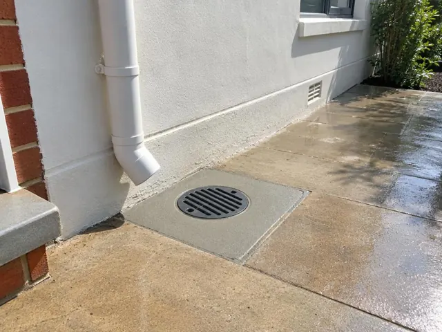 Outdoor drain grate set in concrete paving beside a house wall, with a downpipe and wet pavers in Abermain
